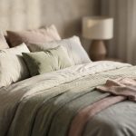Lit luxueux et douillet avec de nombreux coussins et plaids aux textures variées (crème, beige, vert sauge, rose poudré). Lumière douce, ambiance apaisante.